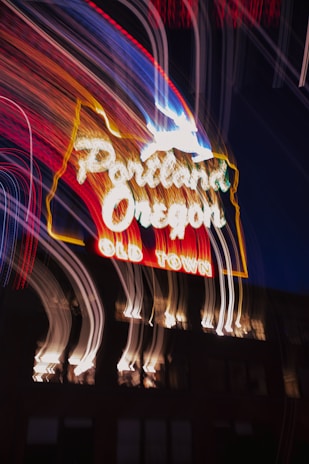 A neon sign with glowing white and red lettering spells out 'Portland, Oregon' and 'Old Town'. Above the text, a white outline of a deer is lit up. The image features motion blur, creating streaks of light across the dark night background, adding a dynamic and vibrant effect.
