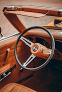 A vintage car interior with a leather-wrapped steering wheel, featuring a classic emblem at its center. The dashboard and upholstery are in a rich brown leather, complemented by chrome accents. The convertible top is down, highlighting the retro design of the vehicle.