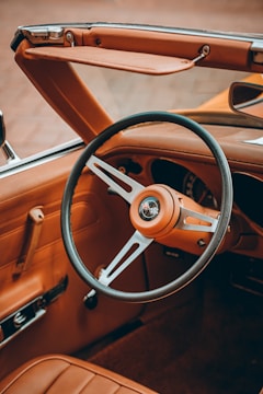 A vintage car interior with a leather-wrapped steering wheel, featuring a classic emblem at its center. The dashboard and upholstery are in a rich brown leather, complemented by chrome accents. The convertible top is down, highlighting the retro design of the vehicle.