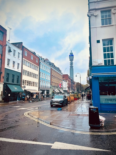 A rainy street scene features a row of colorful buildings, including red, blue, and white facades. A Mercedes car is parked by the curb, and a Union Jack flag is prominently displayed. A few pedestrians walk along the wet pavement under overcast skies, and a distant telecommunications tower is visible.