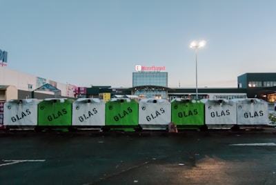 Several recycle bins labeled 'GLAS' are lined up in a row in front of a shopping center. The bins are primarily white and green. The background shows the entrance to a store under a clear sky, with a few commercial signs visible.