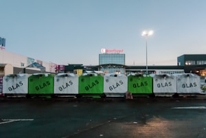 Several recycle bins labeled 'GLAS' are lined up in a row in front of a shopping center. The bins are primarily white and green. The background shows the entrance to a store under a clear sky, with a few commercial signs visible.