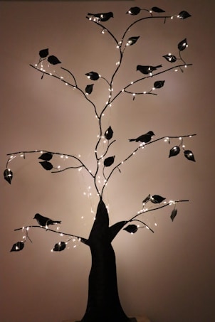 A decorative metal tree sculpture adorned with small string lights. The tree has dark-colored branches with attached leaf shapes and bird silhouettes, casting soft shadows against the wall.