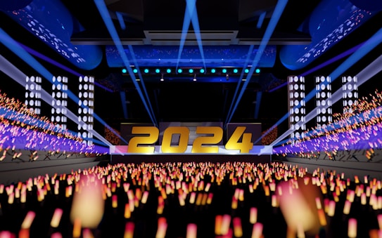 A vibrant and dynamic stage setup featuring large, illuminated numbers '2024' at the center. The foreground shows a crowd of glowing light sticks in orange and pink, creating an energetic atmosphere. The background is filled with bright spotlights and digital screens in a modern indoor venue, giving a sense of celebration and anticipation.