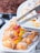 A variety of pastries arranged on a white platter. There are cream puffs dusted with powdered sugar and fruit tarts topped with strawberries, kiwi, and orange slices. A metal tong is placed over a tray of Mille-feuille pastry with chocolate drizzle.