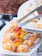 A variety of pastries arranged on a white platter. There are cream puffs dusted with powdered sugar and fruit tarts topped with strawberries, kiwi, and orange slices. A metal tong is placed over a tray of Mille-feuille pastry with chocolate drizzle.