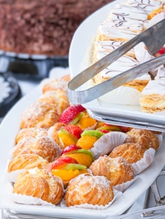 A variety of pastries arranged on a white platter. There are cream puffs dusted with powdered sugar and fruit tarts topped with strawberries, kiwi, and orange slices. A metal tong is placed over a tray of Mille-feuille pastry with chocolate drizzle.