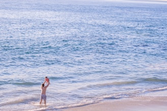 A person stands in shallow ocean water at the beach, lifting a child in the air. The child is wearing a bright outfit, and the water stretches out to the horizon, appearing calm and blue under the sunlight.