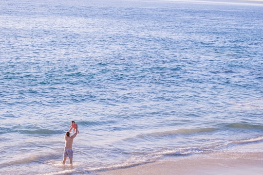 A person stands in shallow ocean water at the beach, lifting a child in the air. The child is wearing a bright outfit, and the water stretches out to the horizon, appearing calm and blue under the sunlight.
