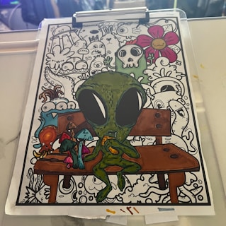 A colorful drawing features a green alien creature sitting on a wooden bench, surrounded by whimsical and abstract doodles. The alien has large black eyes and is holding an orange object. Brightly colored mushrooms and an octopus-like creature are in the foreground, while doodles of various creatures, a skeleton, and a smiling flower fill the background.