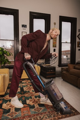 A person wearing a maroon suit is leaning back while vacuuming a rug in a neatly arranged room. The room features large windows with black frames, a plant, and various pieces of furniture such as a yellow armchair and a brown sofa. The walls are adorned with framed pictures and artwork.