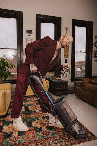 A person wearing a maroon suit is leaning back while vacuuming a rug in a neatly arranged room. The room features large windows with black frames, a plant, and various pieces of furniture such as a yellow armchair and a brown sofa. The walls are adorned with framed pictures and artwork.