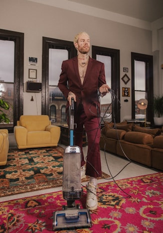 A person with blond hair is dressed in a burgundy suit while using a blue and gray vacuum cleaner in a well-decorated living room. The room features large windows, a mix of colorful rugs, and a variety of furniture pieces including mustard and brown couches. Several pieces of wall art and potted plants are visible, contributing to a cozy and artistic atmosphere.