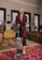 A person with blond hair is dressed in a burgundy suit while using a blue and gray vacuum cleaner in a well-decorated living room. The room features large windows, a mix of colorful rugs, and a variety of furniture pieces including mustard and brown couches. Several pieces of wall art and potted plants are visible, contributing to a cozy and artistic atmosphere.