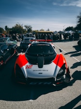 A high-performance sports car with a sleek aerodynamic design is parked in an outdoor setting. It features a prominent red and white paint scheme, with the number 12 and 'Richard Mille' displayed on its front. The background includes other luxury vehicles and a crowd of people, indicating a car show or gathering. The sky is partly cloudy with visible trees in the distance.
