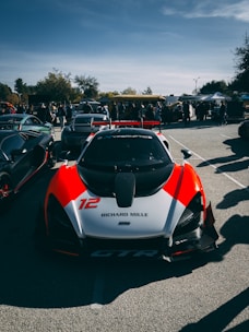 A high-performance sports car with a sleek aerodynamic design is parked in an outdoor setting. It features a prominent red and white paint scheme, with the number 12 and 'Richard Mille' displayed on its front. The background includes other luxury vehicles and a crowd of people, indicating a car show or gathering. The sky is partly cloudy with visible trees in the distance.