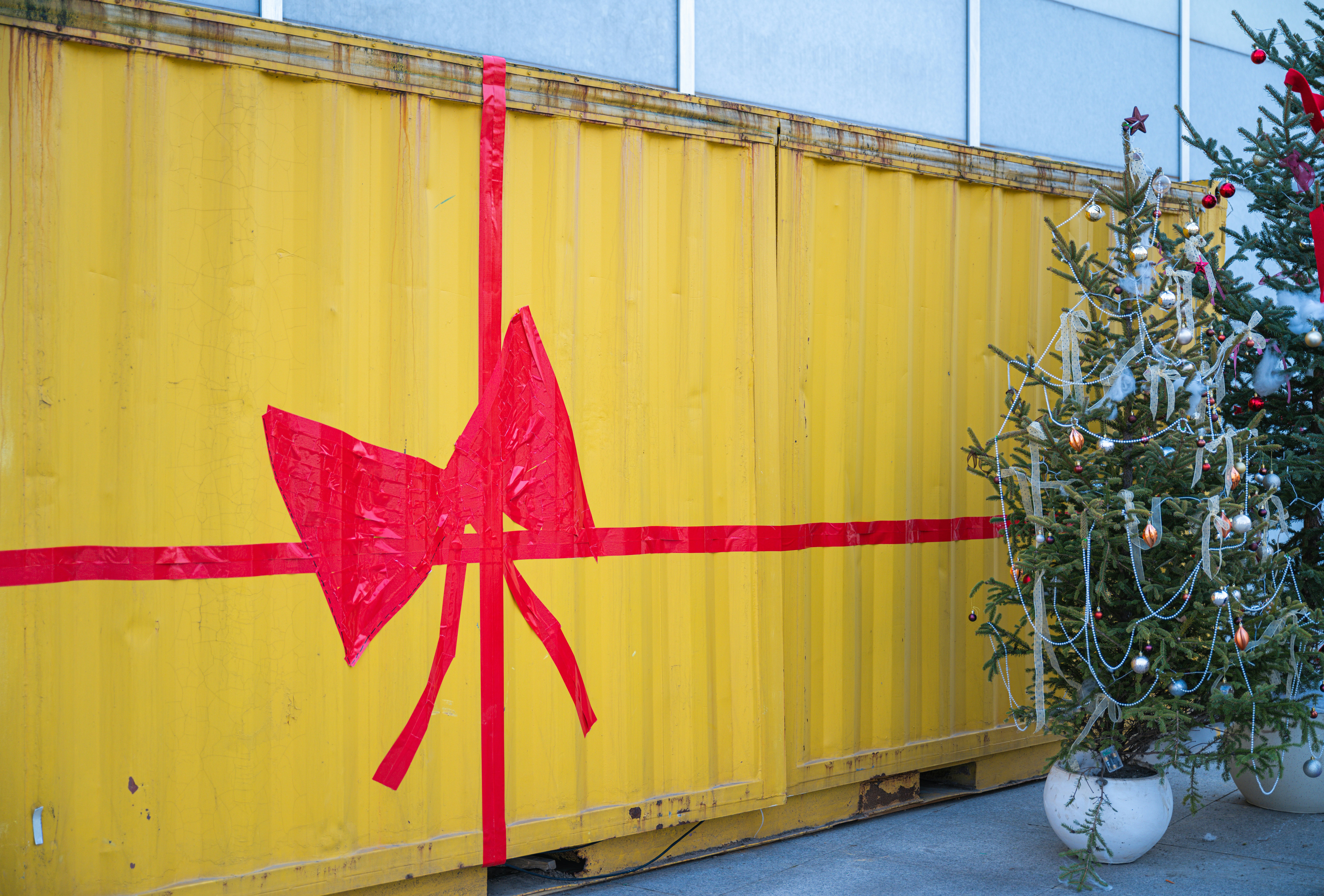 a christmas tree sitting next to a yellow container, Red ribbon pattern on yellow shipping container with two Christmas trees next to it
