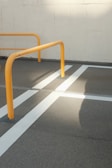 Yellow metal barriers are positioned on a gray concrete surface with crisp white lines creating a geometric pattern. The sunlight casts a distinct shadow across the pavement, highlighting the structure and lines.