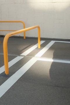 Yellow metal barriers are positioned on a gray concrete surface with crisp white lines creating a geometric pattern. The sunlight casts a distinct shadow across the pavement, highlighting the structure and lines.