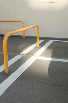 Yellow metal barriers are positioned on a gray concrete surface with crisp white lines creating a geometric pattern. The sunlight casts a distinct shadow across the pavement, highlighting the structure and lines.