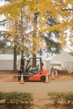 A forklift is parked on a dirt path surrounded by orange traffic cones. In the background, there are several white tents and tall trees with vibrant yellow foliage, indicative of autumn. The ground is covered with fallen leaves, adding to the autumnal atmosphere.