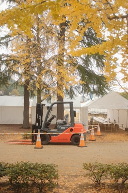 A forklift is parked on a dirt path surrounded by orange traffic cones. In the background, there are several white tents and tall trees with vibrant yellow foliage, indicative of autumn. The ground is covered with fallen leaves, adding to the autumnal atmosphere.