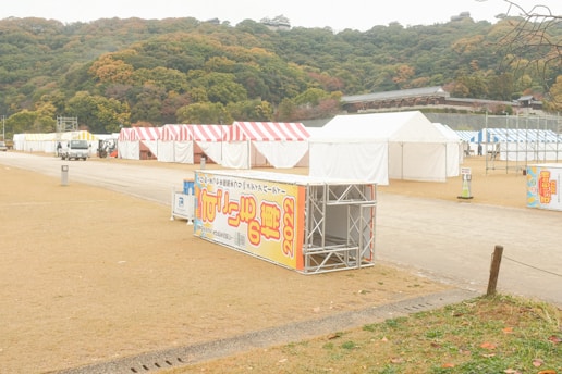 Several tents with striped red, yellow, and white covers are set up in an open area with a slight hill in the background. A few vehicles are present, and there are large signs with text and the year 2022. The surrounding trees have autumn colors, suggesting a seasonal event.