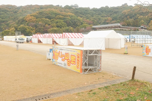 Several tents with striped red, yellow, and white covers are set up in an open area with a slight hill in the background. A few vehicles are present, and there are large signs with text and the year 2022. The surrounding trees have autumn colors, suggesting a seasonal event.