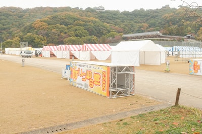 Several tents with striped red, yellow, and white covers are set up in an open area with a slight hill in the background. A few vehicles are present, and there are large signs with text and the year 2022. The surrounding trees have autumn colors, suggesting a seasonal event.