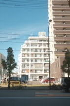 A residential urban setting features a white mid-rise apartment building with balconies in the center. Adjacent is a taller brown building with numerous balconies. Several cars are parked in a lot in front of the buildings, while trees and a street with visible road markings occupy the foreground. Overhead wires and a streetlamp are also visible.