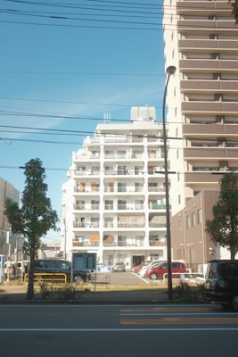 A residential urban setting features a white mid-rise apartment building with balconies in the center. Adjacent is a taller brown building with numerous balconies. Several cars are parked in a lot in front of the buildings, while trees and a street with visible road markings occupy the foreground. Overhead wires and a streetlamp are also visible.