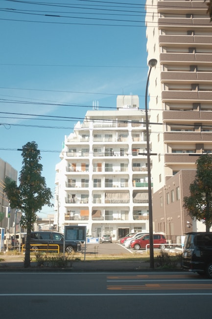 A residential urban setting features a white mid-rise apartment building with balconies in the center. Adjacent is a taller brown building with numerous balconies. Several cars are parked in a lot in front of the buildings, while trees and a street with visible road markings occupy the foreground. Overhead wires and a streetlamp are also visible.