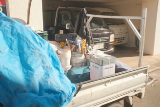 A pickup truck with various tools and supplies in its bed is parked in a driveway. Items include buckets, a toolbox, a blue tarp, and a metal container labeled 'kikusui'. In the background, two vehicles are parked in a garage.