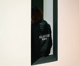 A person wearing a dark jacket with 'VOLUNTEER' and some Chinese characters written on the back is standing partially hidden in an architectural opening. The background is mostly dark, creating a contrast with the lighter frame of the wall.
