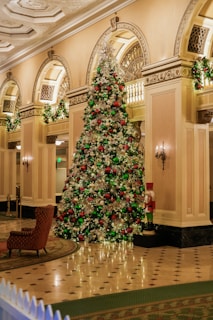 A grand and elaborately decorated Christmas tree stands in the center of an opulent hotel lobby, adorned with silver, red, and green ornaments, as well as twinkling lights. The surrounding architecture features ornate columns, arches, and intricate moldings. A comfortable armchair sits nearby, and a decorative nutcracker figure is positioned next to the tree.