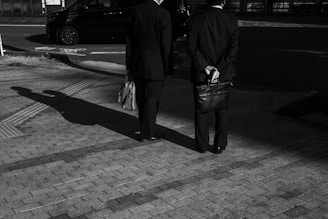 Two men in suits stand on a paved surface under the streetlights, casting long shadows. One holds a briefcase behind his back while the other carries a bag. A car is parked on the street in the background.