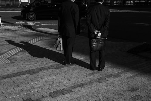 Two men in suits stand on a paved surface under the streetlights, casting long shadows. One holds a briefcase behind his back while the other carries a bag. A car is parked on the street in the background.