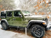 A green, four-door Jeep with the model name 'WILLYS' on the hood is parked outdoors during a snowfall. There are trees in the background with some branches covered in snow, while others have dry leaves.