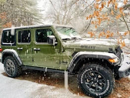 A green, four-door Jeep with the model name 'WILLYS' on the hood is parked outdoors during a snowfall. There are trees in the background with some branches covered in snow, while others have dry leaves.