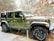 A green, four-door Jeep with the model name 'WILLYS' on the hood is parked outdoors during a snowfall. There are trees in the background with some branches covered in snow, while others have dry leaves.