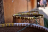 A row of woven baskets with green rope handles is positioned against a blurred background including a wall and out-of-focus figures. The baskets are made from natural materials, featuring intricate patterns and a warm color palette.