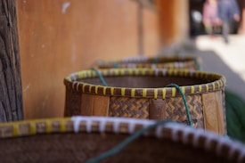 A row of woven baskets with green rope handles is positioned against a blurred background including a wall and out-of-focus figures. The baskets are made from natural materials, featuring intricate patterns and a warm color palette.