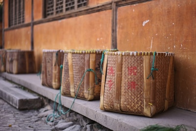 A row of traditional woven baskets with handles made of rope sits on a stone platform against an orange wall. The baskets have red inscriptions on them and appear to be made of bamboo or similar material.