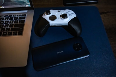 A white and black game controller rests on a blue surface alongside a Nokia smartphone positioned near a laptop. The laptop has a visible keyboard and screen, indicating it's powered on.