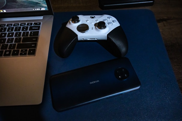 A white and black game controller rests on a blue surface alongside a Nokia smartphone positioned near a laptop. The laptop has a visible keyboard and screen, indicating it's powered on.