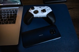 A white and black game controller rests on a blue surface alongside a Nokia smartphone positioned near a laptop. The laptop has a visible keyboard and screen, indicating it's powered on.
