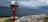A reflective sculpture of a figure painted red is holding a large, shiny sphere that reflects the surrounding landscape. The sculpture is set near a rocky seaside cliff with a view of the ocean. The sky is cloudy, with varying shades of blue and grey.
