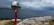 A reflective sculpture of a figure painted red is holding a large, shiny sphere that reflects the surrounding landscape. The sculpture is set near a rocky seaside cliff with a view of the ocean. The sky is cloudy, with varying shades of blue and grey.