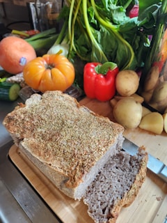 A freshly baked loaf of bread topped with herbs is placed on a wooden cutting board, alongside a knife with a shiny blade. In the background, there is an assortment of colorful vegetables, including a large tomato, red bell pepper, potatoes, leafy greens, and a squash.