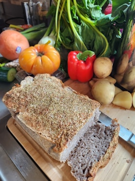 A freshly baked loaf of bread topped with herbs is placed on a wooden cutting board, alongside a knife with a shiny blade. In the background, there is an assortment of colorful vegetables, including a large tomato, red bell pepper, potatoes, leafy greens, and a squash.